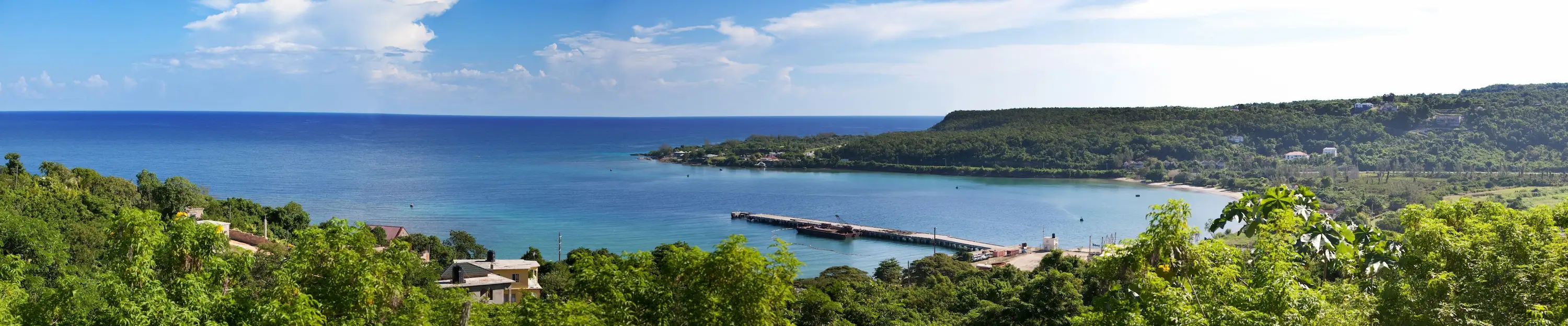 Panoramic view of a turquoise Caribbean bay with a pier, surrounded by lush green hills under a blue sky.